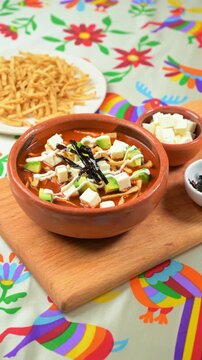 Close-up of Aztec soup with fresh ingredients, cubed cheese and fried strips of pasilla chile, served on a table with a Mexican tablecloth