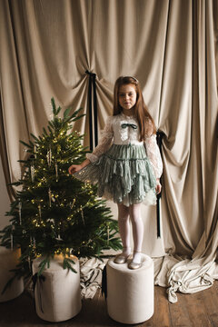 Smiling child girl 5-6 year old wearing princess dress decorating Christmas tree over beige background. Winter holiday season.
