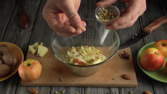 Hands of a person adding spices to a bowl of chopped apples on a wooden cutting board with nuts and apples arranged around the scene