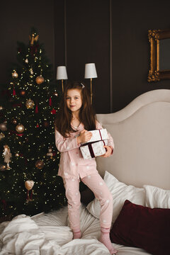 Funny child girl 5-6 year old holding Christmas present box in bed over xmas tree in bedroom. Winter holiday season.