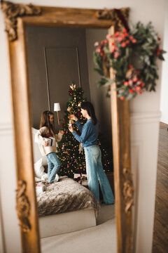 Mother with child girl decorating Christmas tree in bedroom mirror reflection. Winter holiday season. Cozy home atmosphere.