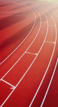 A sweeping aerial view of a vibrant red synthetic athletic track with white lane markings curving gracefully under soft morning sunlight, texture, bend, track