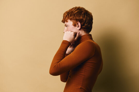 Profile of redhead woman with short curly hair wearing brown turtleneck, standing with closed fists near face against beige background indoor.