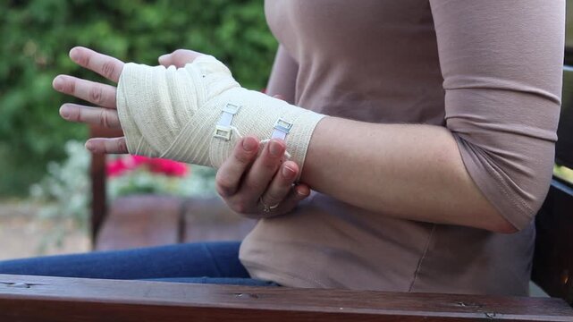 Woman with bandaged hand sitting on bench. Close-up of female hands with elastic bandage on wrist. Hand injury, sprain. Woman holding injured hand bandaged with medical bandage