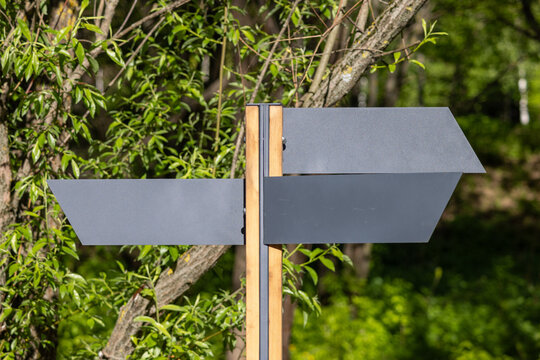 Blank dark gray directional sign with three arrow-shaped panels mounted on a vertical wooden post. Green leaves and a tree trunk fill the sunlit background