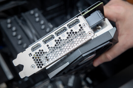 A technician's hand holds a powerful graphics card above a motherboard during gaming PC assembly. The silver rear bracket has three DisplayPort ports and one HDMI port