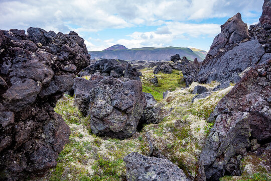 Rugged lava rock formations in icelandic landscape featuring moss-covered terrain and distant mountains under a partly cloudy sky on a bright day with clear visibility.