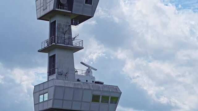 Rotating radar gear and control rooms monitor air and sea traffic from a modern concrete telecom tower beneath a cloudy sky