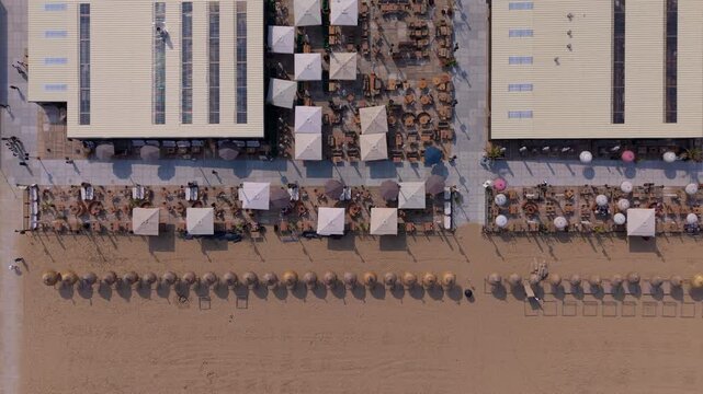 Aerial top down look view of beachfront restaurant complexes with elegant white parasols and a long straight row of thatched beach umbrellas on sand under a bright summer sky