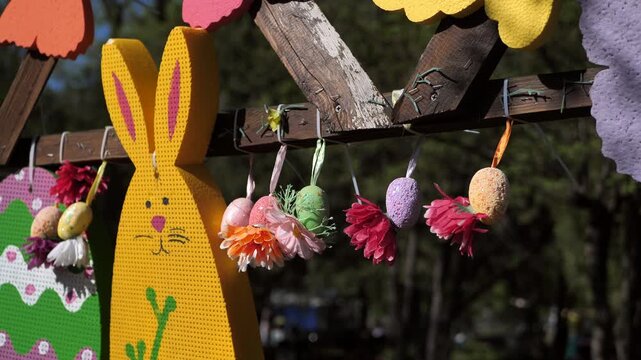 Easter outdoor decorations featuring a yellow wooden rabbit shape and colorful hanging decorative eggs with flowers on a sunny day.