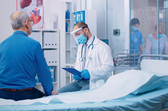 Doctor wearing protective equipment speaks with a senior male patient during a medical consultation in a private clinic or hospital office.