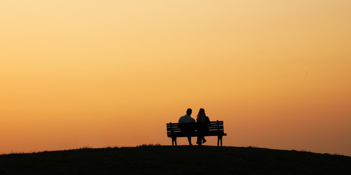 black silhouette of Young adult romantic senior pensioner couple talking kissing on bench on yellow orange sunset sky background backdrop first evening date. unrecognizable person  people