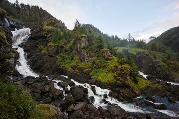 Obraz premium A horizontal scenic shot of a powerful waterfall cascading down rocky mountain cliffs into a rushing river, surrounded by green mossy rocks and pine trees.
