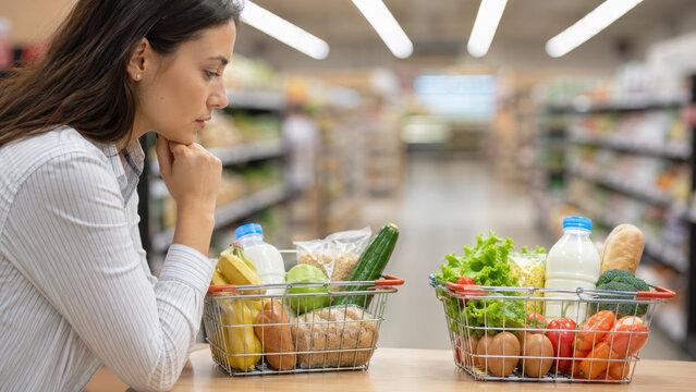 Woman considers her budget while reviewing grocery list with fresh produce dairy and bread in baskets at supermarket