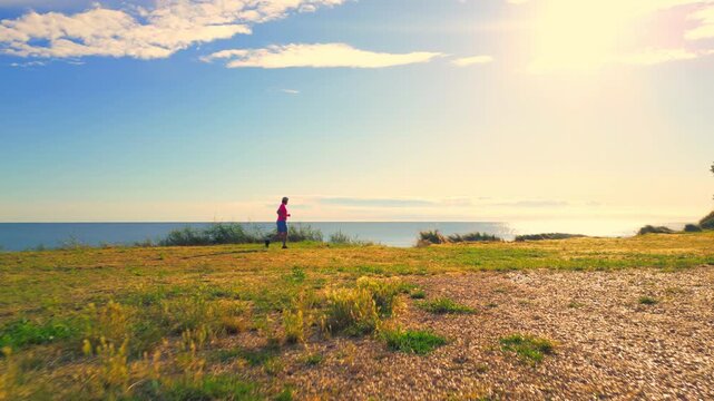 A senior adult is jogging along a grassy cliffside path by the ocean, with the sun reflecting off the water. The serene coastal environment highlights a healthy lifestyle through regular exercise.