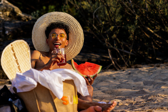 Young man enjoying summer vacation on beach, eating watermelon and drinking cocktail