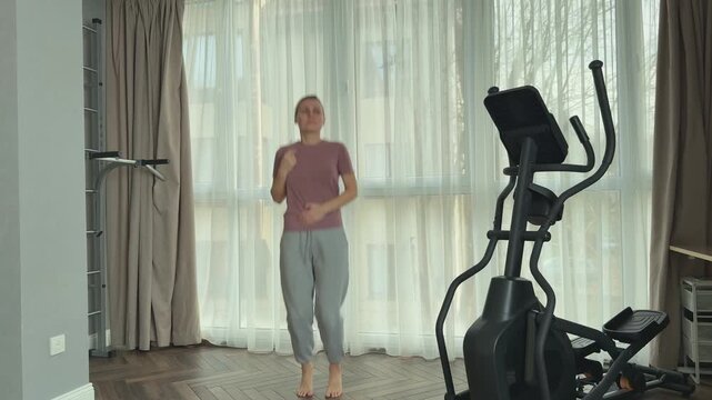 Woman in a pink t shirt, light sports pants and gray sneakers performs jumps in place in a spacious living room with elliptical machine, neutral curtains and wooden floor.