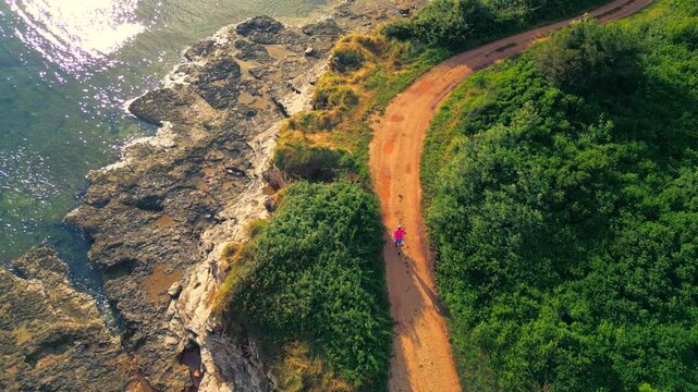 A senior adult jogs along a scenic coastal path surrounded by lush greenery and rocky cliffs. The aerial shot captures the solitary figure in vibrant attire, emphasizing.