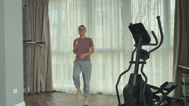Woman in a pink t shirt, light sports pants and gray sneakers stands ready for exercise in a modern living room with elliptical trainer, wooden floor and soft neutral curtains.