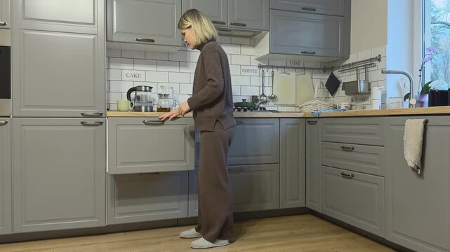 Female puts colander into upper cabinet and pauses thoughtfully in kitchen.
