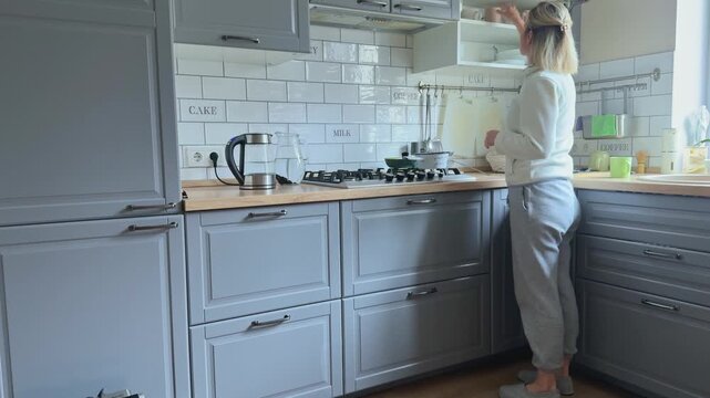 Woman in a light sweatshirt and soft home pants stands in a gray modern kitchen, places cups into the upper cabinet and closes the door. Bright countertop, white tile backsplash and calm domestic 