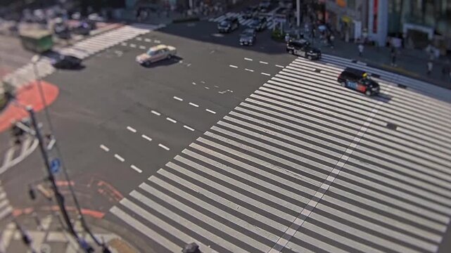 High angle tilt shift timelapse of a busy Tokyo intersection with cars, scooters, and pedestrians crossing wide zebra crosswalks in daylight.