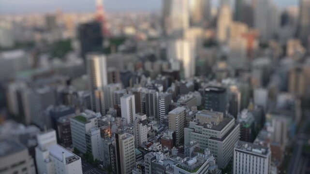 Tilt shift timelapse view over dense Tokyo buildings, shifting from daylight to illuminated night cityscape. Urban skyline with miniature effect and bokeh lights.