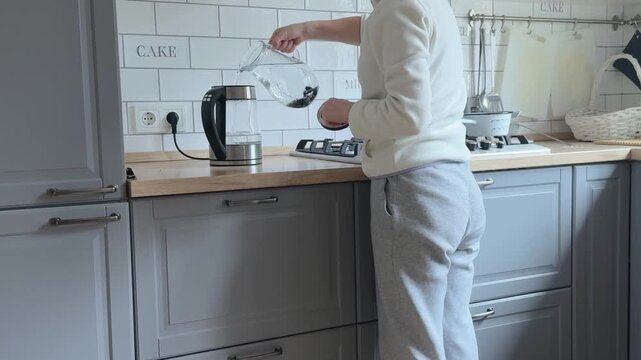Woman in a light sweatshirt and soft pants pours water from a glass carafe into an electric kettle in a modern gray kitchen with white tiled backsplash and wooden countertop. Calm everyday kitchen
