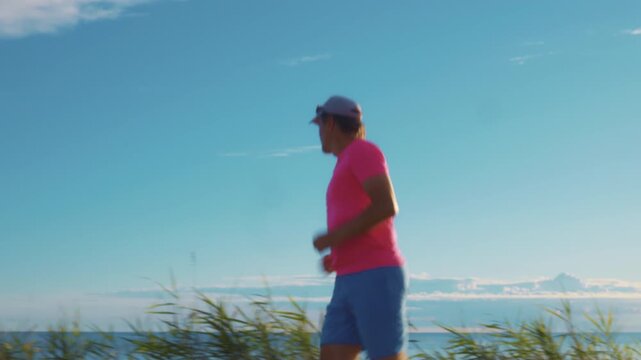 A senior adult man in a pink shirt and blue shorts jogs along a serene coastal path with a clear sky and calm ocean in the background.