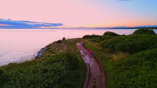 A senior adult jogs along a serene coastal path during sunrise, surrounded by lush greenery and calm waters. The scene promotes a healthy lifestyle through regular exercise and sport activity.