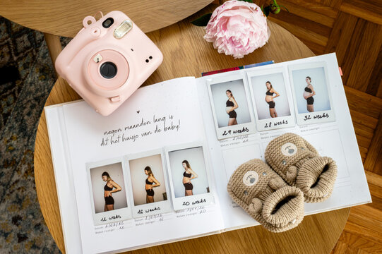 An open pregnancy album with instant photos of the bump by week sits beside a pink instant camera a peony flower and knitted booties a soft maternity flat lay.