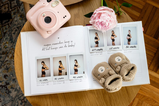 A pregnancy journal filled with instant photos of the growing belly lies open with knitted baby booties a pink peony and an instant camera a tender maternity flat lay.
