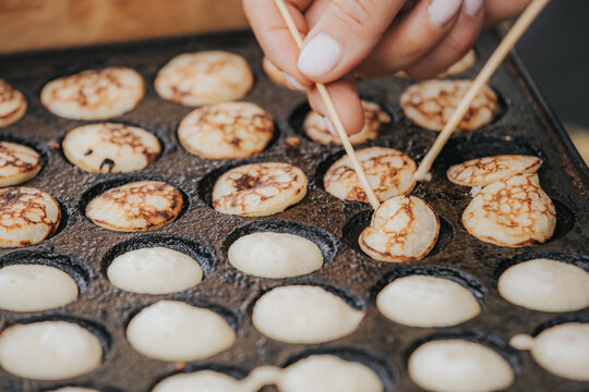 Close-up of a hand using chopsticks to remove round pancakes from a traditional cast iron pan with multiple circular molds on a wooden surface