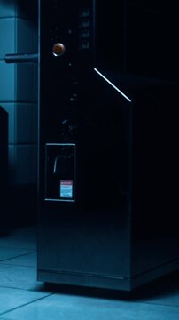A vending machine is positioned in a dimly lit area of a building. Its design is simple and metallic. The tiles on the wall behind it are white. Light from the room highlights the machines features.