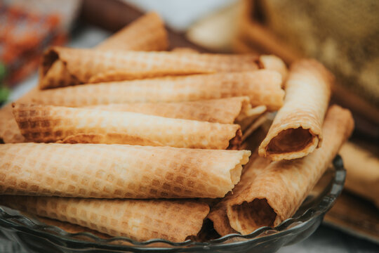 Stack of crispy wafer rolls arranged in a glass bowl on a textured surface with blurred background elements suggesting a cozy kitchen setting