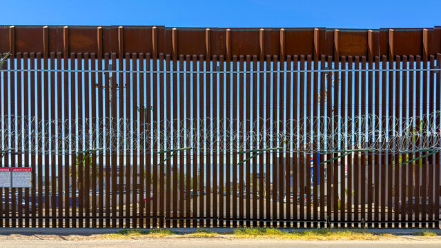 Straight-on view of the tall metal US-Mexico border wall with razor wire between Calexico and Mexicali under a clear blue sky