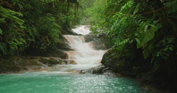 Tiered Blue Falls cascade in Costa Rica flowing into turquoise pool with visitors on hanging bridge