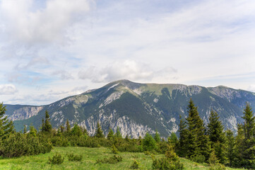 Obraz premium A breathtaking view of Rax Mountain in Austria featuring rolling green hills and rugged peaks under a vibrant cloudy sky with soft sunlight illuminating the lush greenery