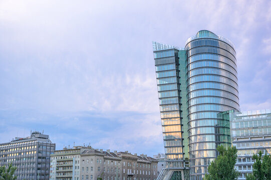 A striking modern building in Austria features sleek glass panels reflecting the soft blue sky with hints of clouds