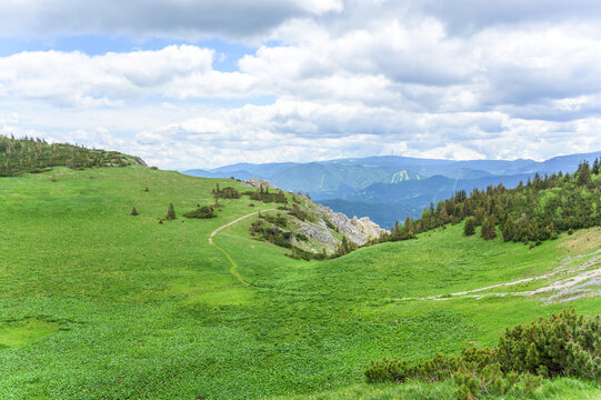 The image showcases a breathtaking mountain landscape in Rax, Austria, dominated by lush green hills and a bright blue sky adorned with fluffy white clouds, creating a stunning contrast with the viv