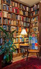 Cozy home library reading corner with floor-to-ceiling wooden bookshelves, a glowing lamp, and a wooden chair with a colorful patchwork quilt © Vit Onopriienko