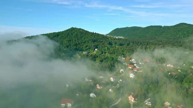 Aerial view of a serene village nestled in a lush forest, with clouds gently rolling over the landscape, creating a peaceful and tranquil scenery.