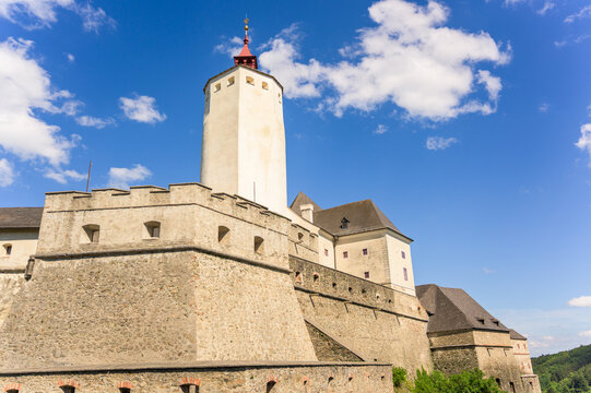 Esterhazy Castle showcases its majestic stone walls and tall turret against a brilliant blue sky filled with scattered clouds, while the surrounding landscape features gentle hills and lush greenery