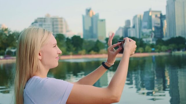 A blonde woman takes a photo of a cozy park lake with fountains, set against the skyscrapers of Bangkok's business district during daytime.