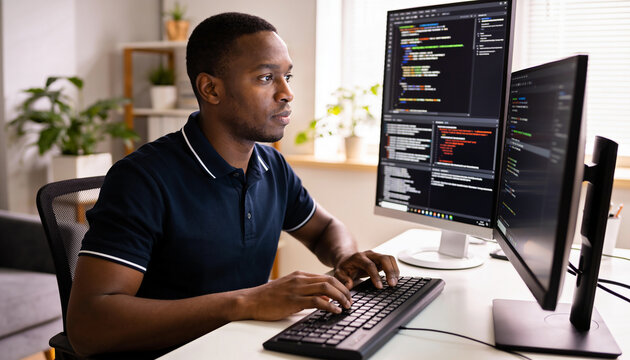 Focused black software developer typing computer code on dual monitors in modern home office with natural light