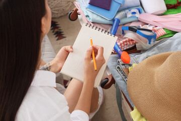 Woman writing a packing list in a notebook with a pencil, preparing for a summer vacation trip with a suitcase full of clothes and travel accessories © Pixel-Shot