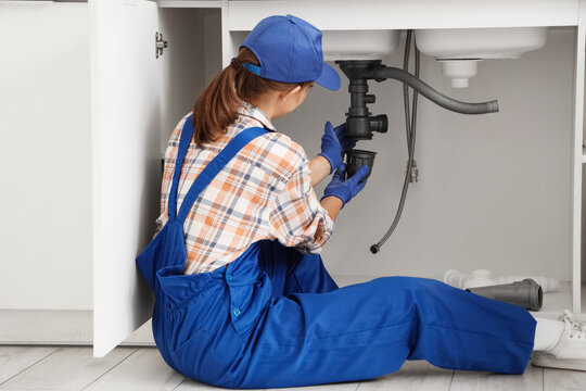 Female plumber in blue uniform and cap repairing a kitchen sink drain pipe under the cabinet, She is sitting on the floor under the cabinet, wearing gloves and focused on the plumbing system