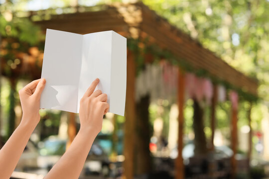 Hands holding a blank white folded brochure or leaflet outdoors, pointing to empty space for copy or design