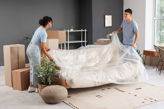 Young couple covering sofa with film in room on moving day