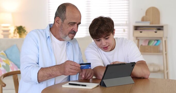 Caregiver. Teenager showing senior man how to pay online with tablet and credit card at wooden table indoors